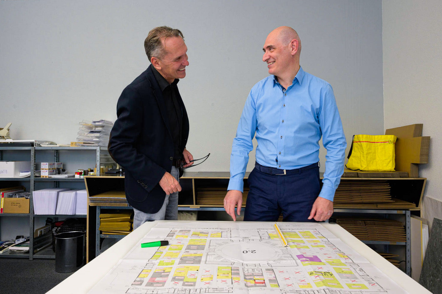 Zwei Männer stehen lächelnd an einem Tisch mit einem grossen Architekturplan. Der Raum enthält Regale mit Ordnern und Dokumenten, auf dem Tisch liegt ein Stift und ein Textmarker.