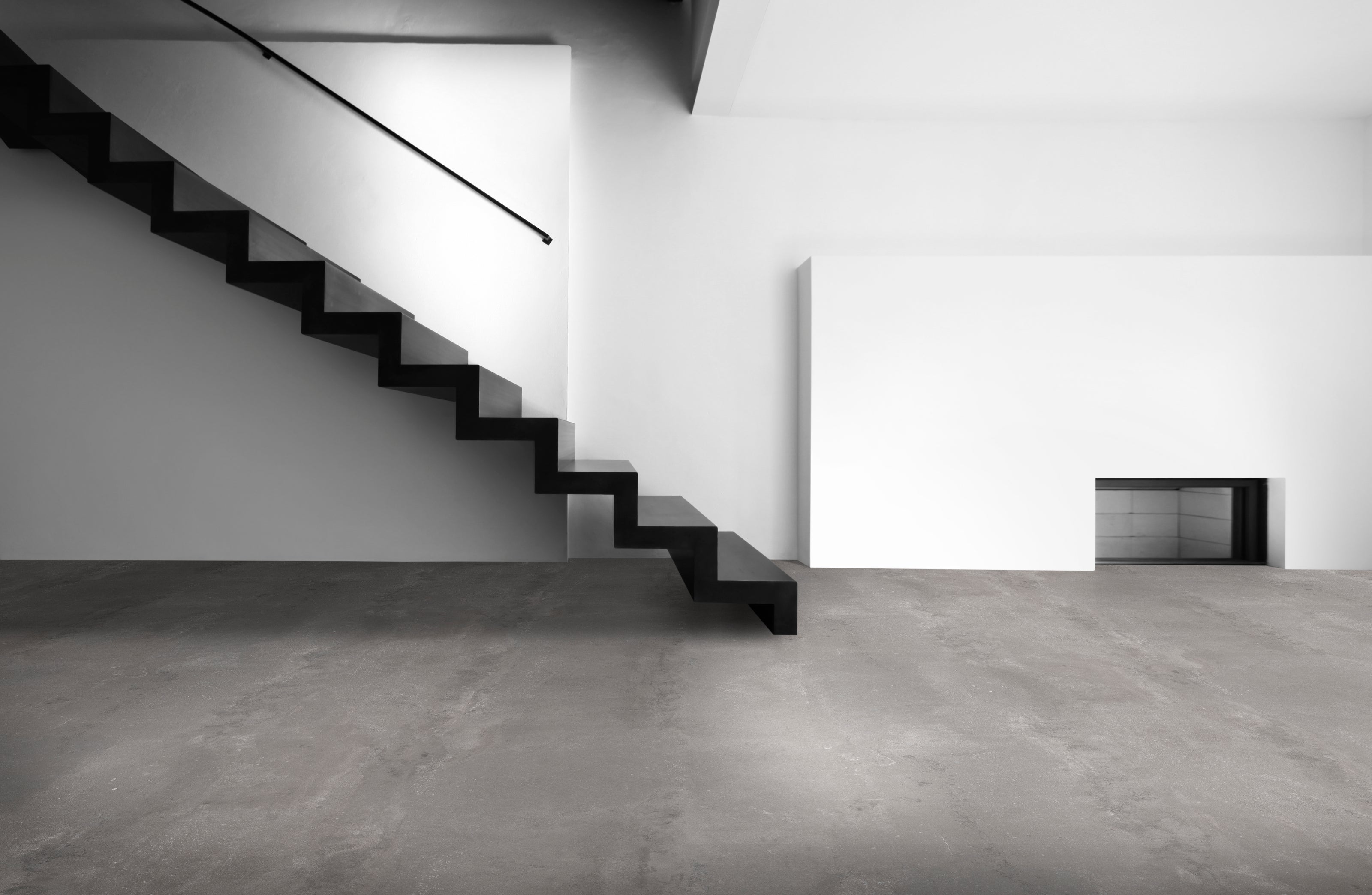 Eine freischwebende, schwarze Treppe mit schlichtem Geländer verläuft seitlich an einer weißen Wand. Der Boden besteht aus glattem, grauem Beton. Minimalistisches Design dominiert.