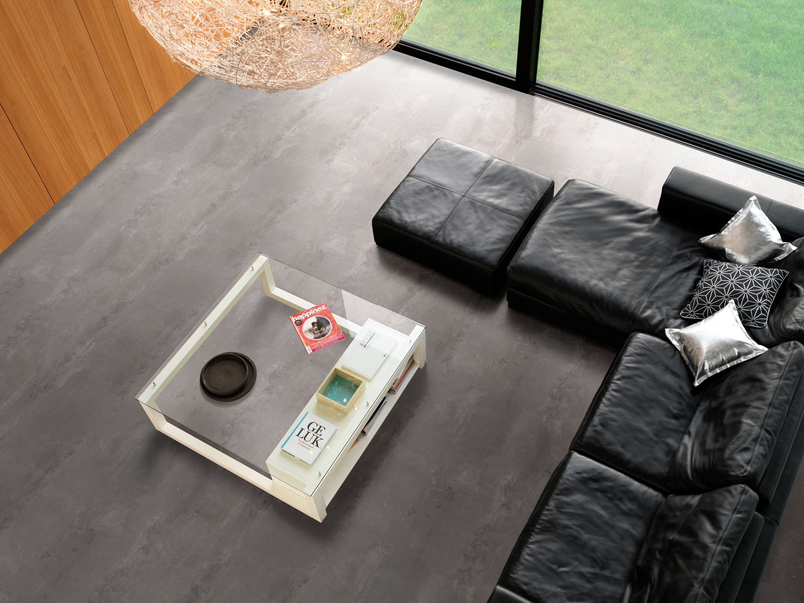 Wohnzimmer mit grauem PVC-Boden in Beton-Optik, schwarzem Ledersofa, Glas-Couchtisch und modernen Dekorationen; große Fenster bieten Blick auf grünen Rasen. Bücher und Zeitschriften liegen auf dem Tisch.