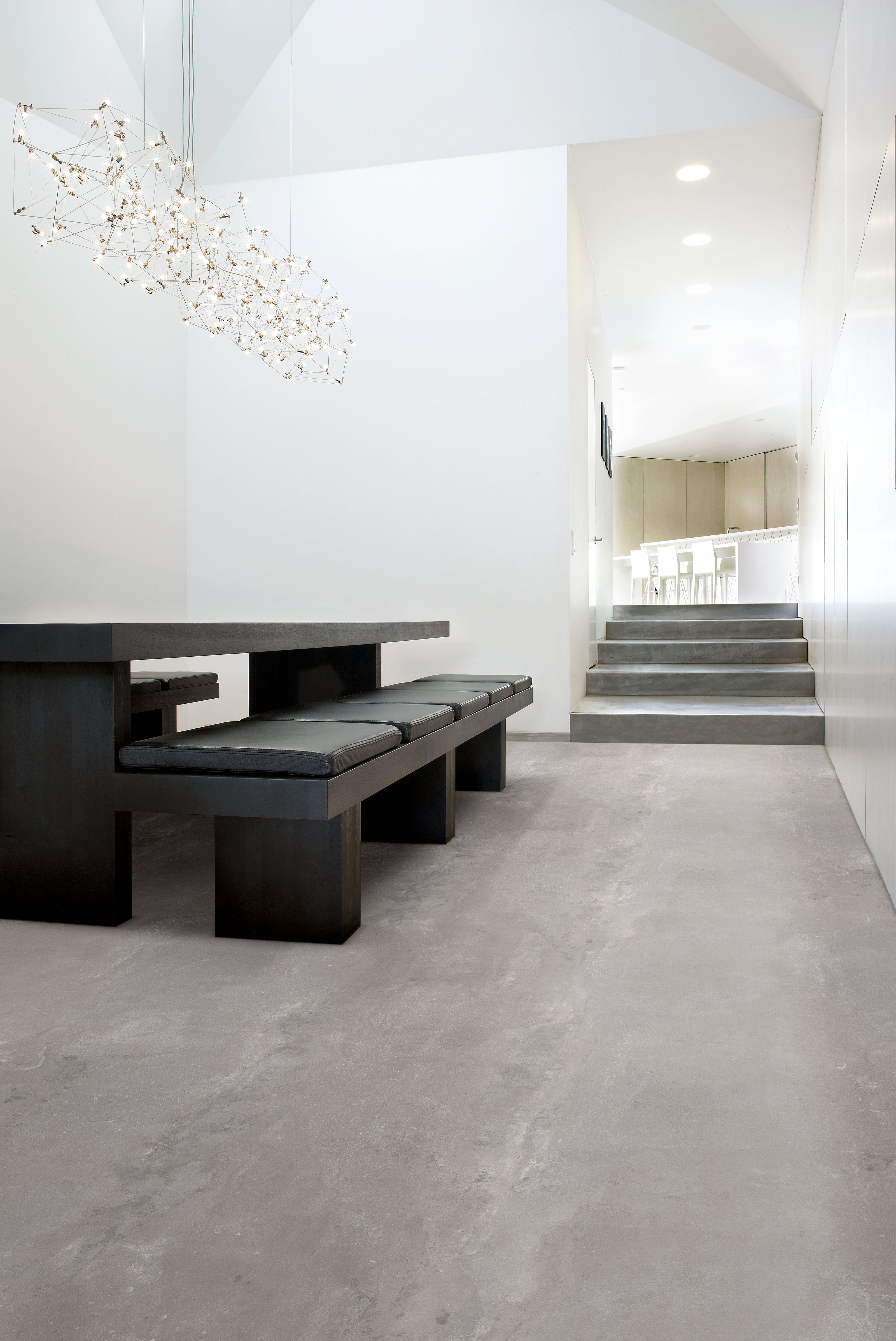 Moderne Esszimmerbank und Tisch vor grauem, glänzendem Boden. Stufen führen zu einem hellen, minimalistischen Küchenbereich. Eine geometrische Lampe hängt von der Decke.