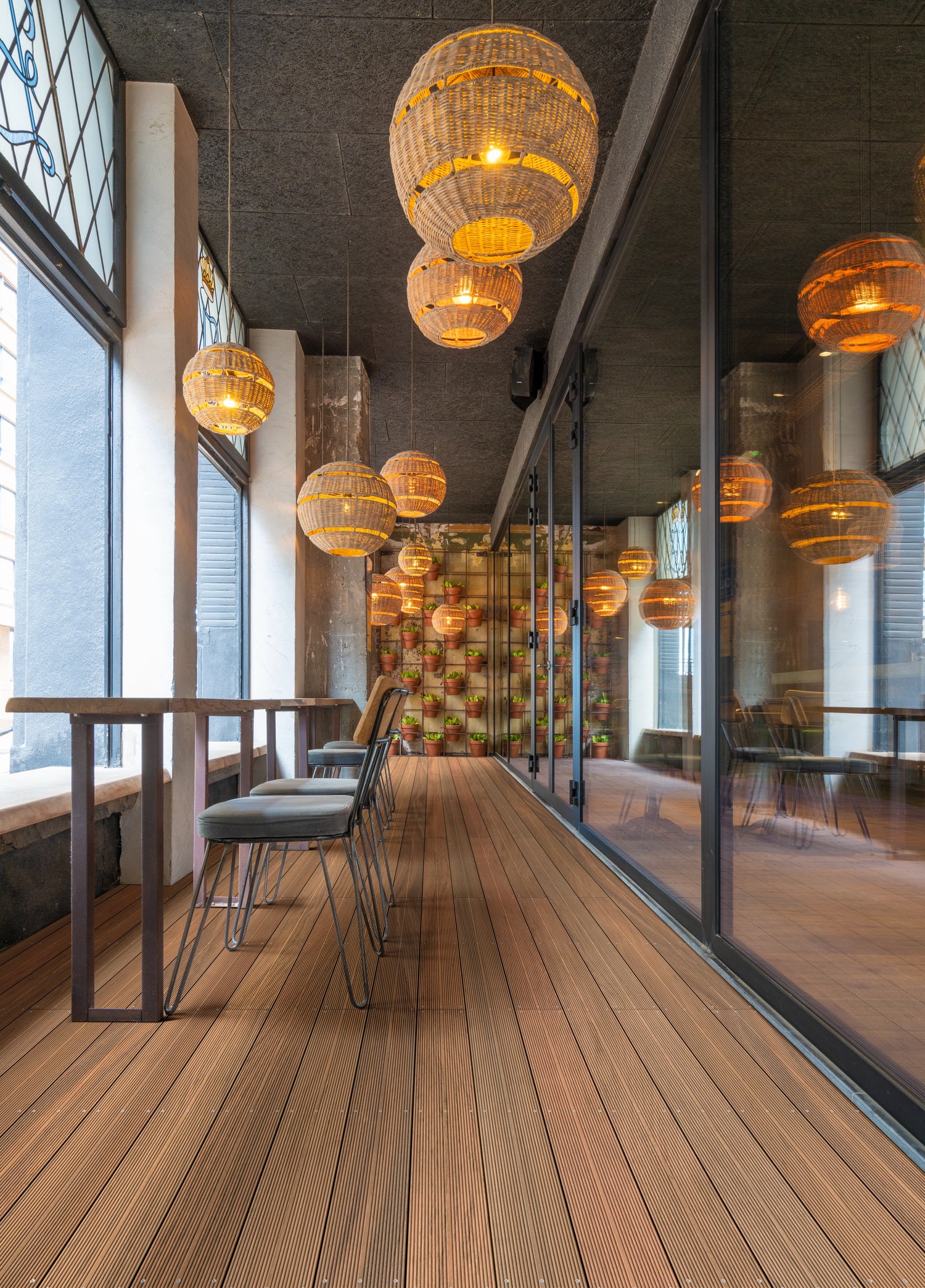 Holzboden-Patio mit Barhockern an einem hohen Tisch; dekorative, beleuchtete Körbe hängen von der Decke. Eine Glaswand reflektiert das gemütliche Ambiente mit Topfpflanzen im Hintergrund.