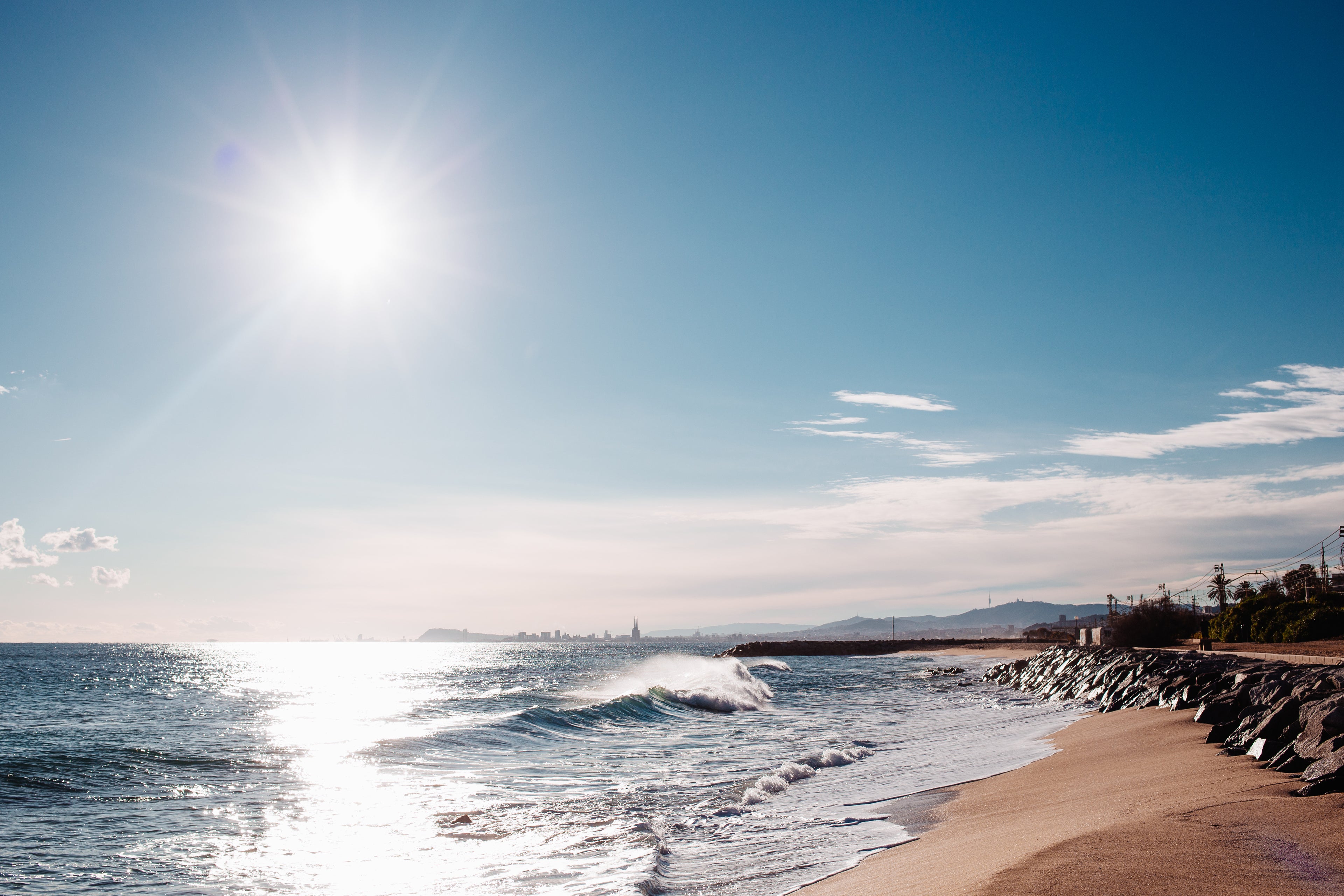 Sonniges Strandpanorama mit strahlender Sonne, Wellen, die auf den Sand treffen, Küstenlinie mit Felsen und entfernter Stadtsilhouette im Hintergrund.