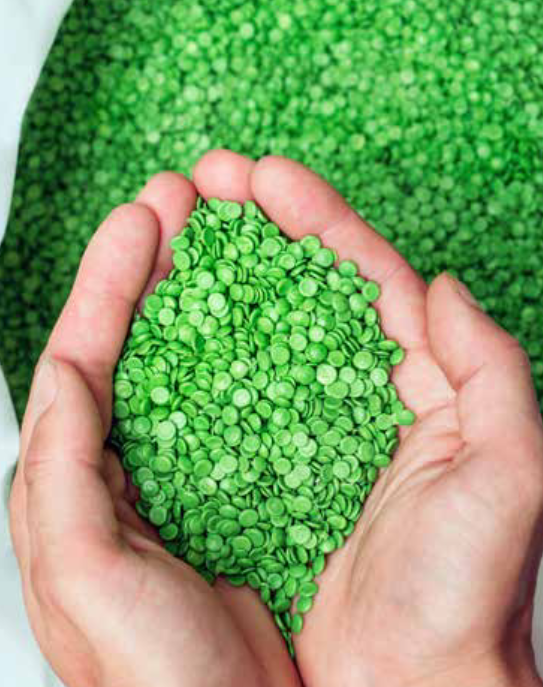 Hände halten eine Menge grüner Kunststoffpellets, aus denen rezyklierte Teppiche hergestellt werden. Im Hintergrund liegt eine grössere Menge derselben Pellets in einem Behälter.